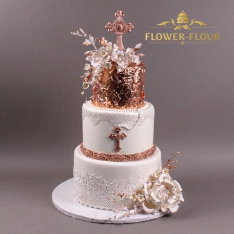 A three-tier baptism cake with a rose-gold top tier, lace, a cross, and white sugar flowers