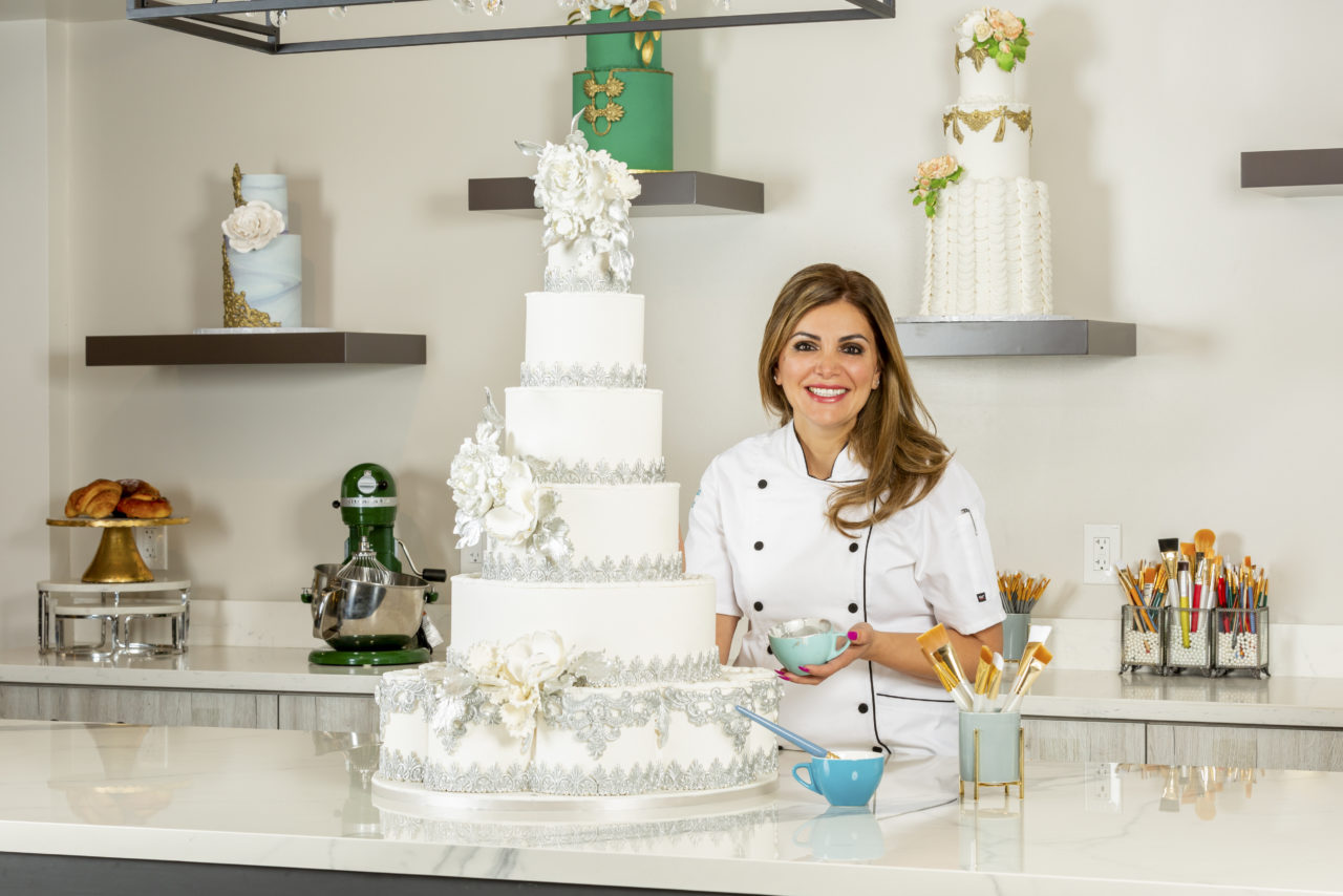 Ani in a white chef’s coat, smiling beside a tall white tiered cake with silver lace and sugar flowers in her studio