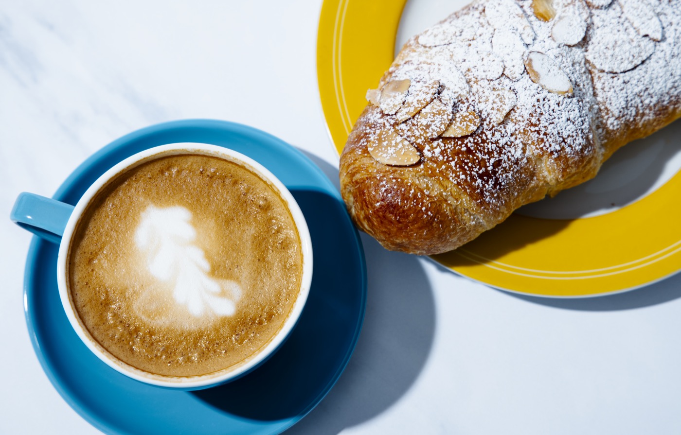 An almond croissant dusted with powdered sugar and sliced almonds on a yellow plate, beside a latte in a blue cup