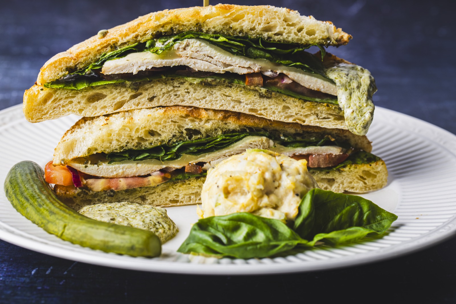 A stacked focaccia sandwich with chicken, greens, and pesto, served with a pickle and basil