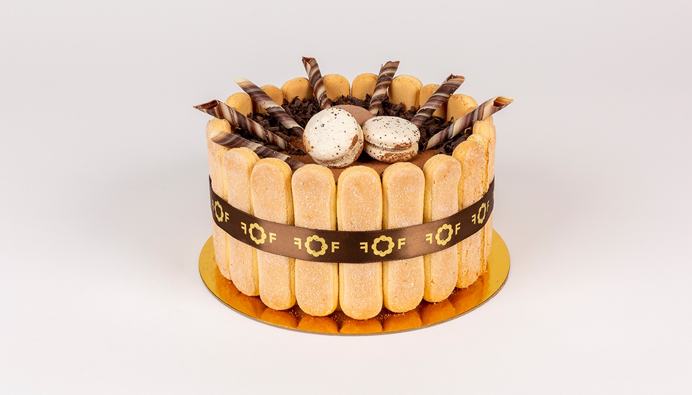 A round tiramisu ringed with ladyfingers and a Flower Flour ribbon, topped with chocolate shavings, macarons, and striped chocolate sticks