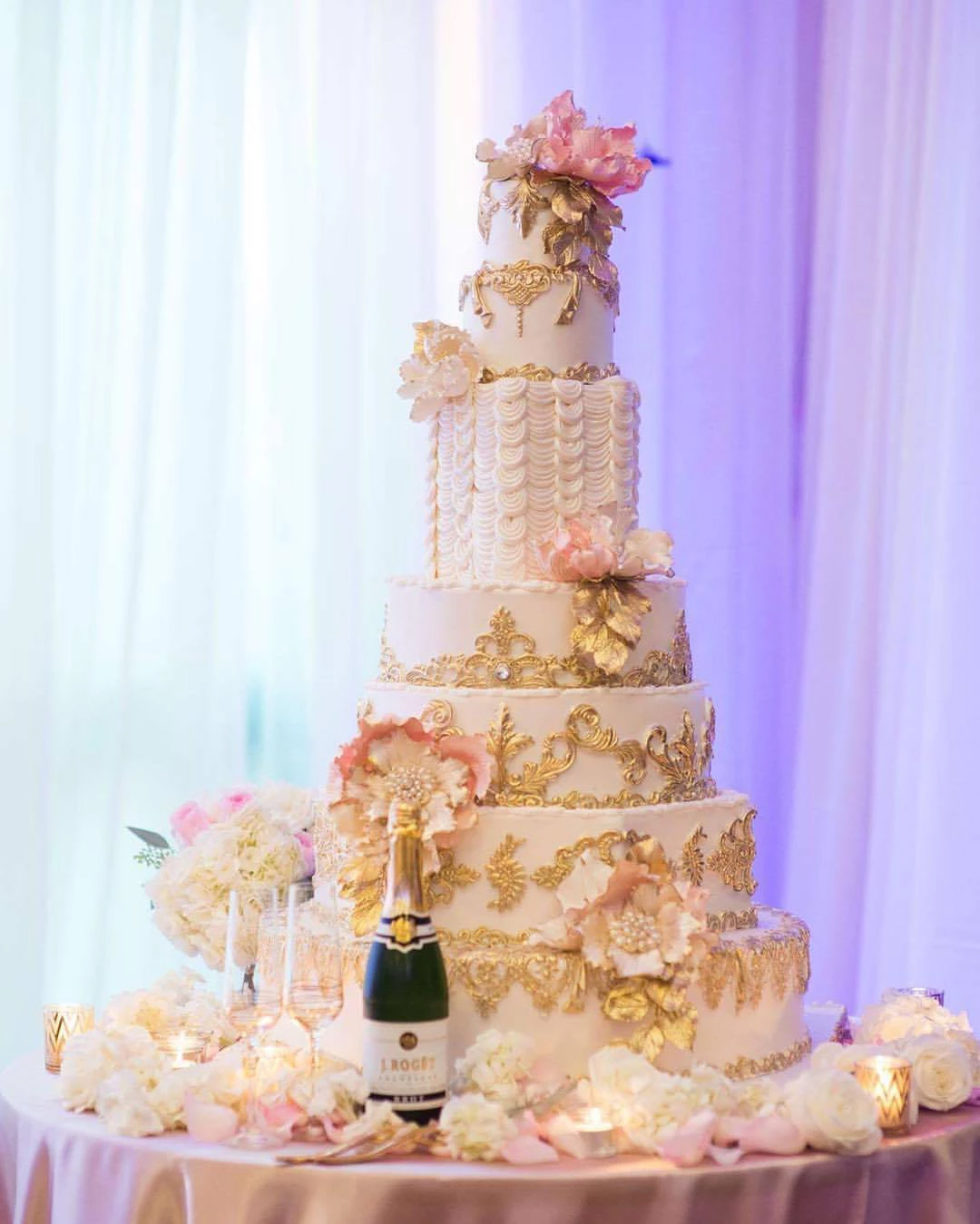 A tall ivory wedding cake with gold baroque detailing and blush sugar peonies on a table of white roses